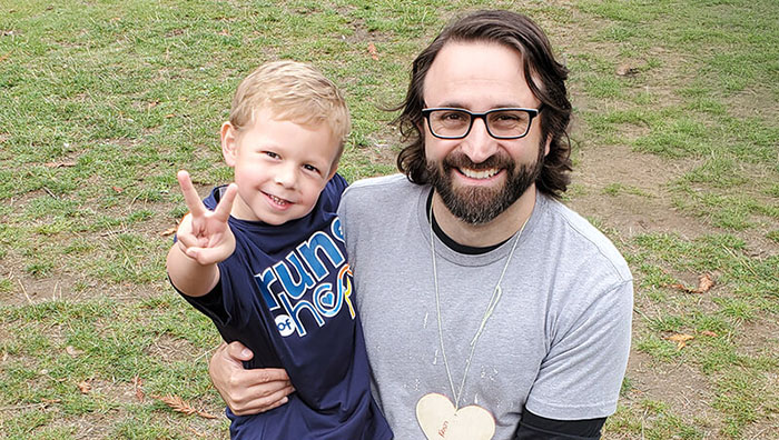 A man with dark hair and glasses is smiling while holding a young boy with blond hair who is showing a peace sign with his fingers and also smiling..