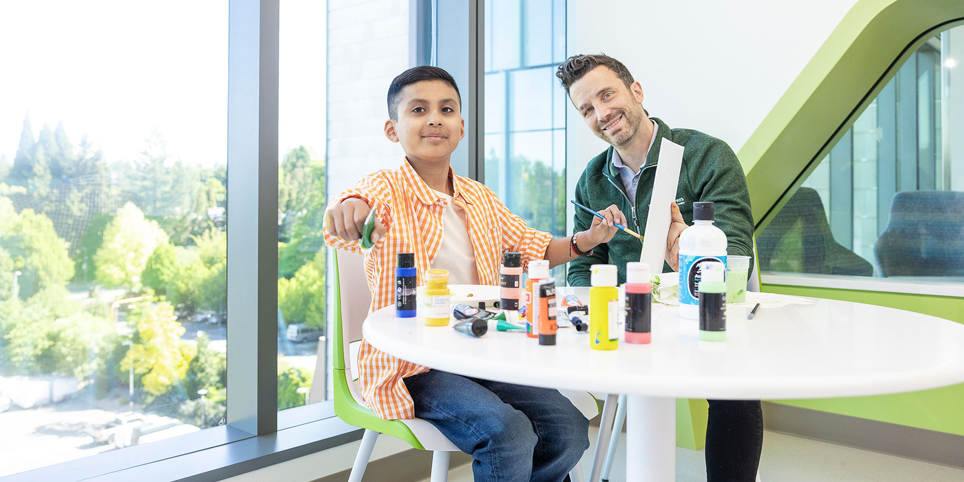 Ino, a young boy, points at the camera. In front of him is a table covered with paints and art supplies. Mike, his art therapist, sits next to him and smiles at the camera while holding a canvas for Ino to paint on.